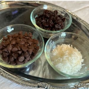 Photo of chocolate chips, dried cherries, and coconut flakes, each in their ramekins, sitting on a silver tray to showcase ingredients used in the brownie recipe.