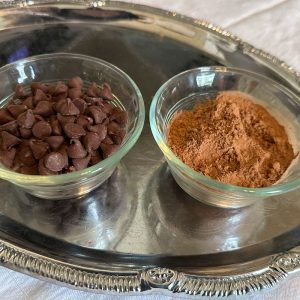 Photo of chocolate chips and cocoa powder, each in their ramekins, sitting on a silver tray to showcase ingredients used in the brownie recipe.